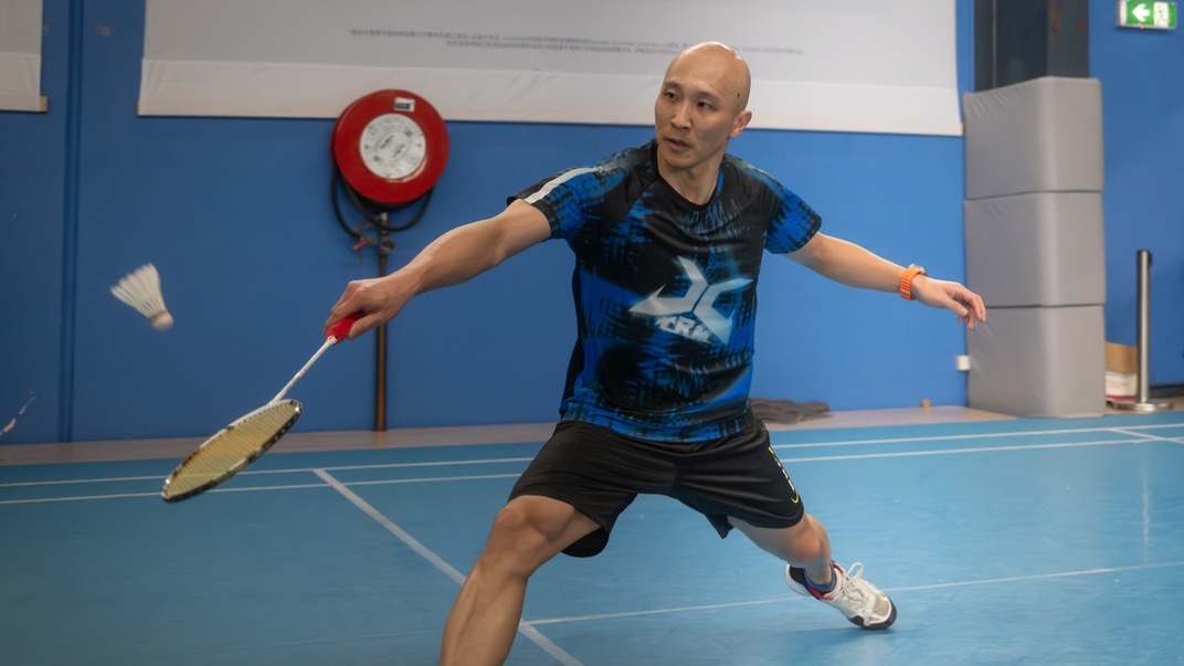 Joe Wu lunging forward to reach a shuttle with his racquet on an indoor badminton court.